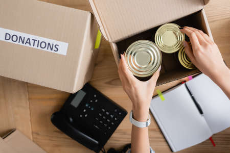 Top view of woman holding tin cans near boxes with donations lettering, telephone and notebook on...