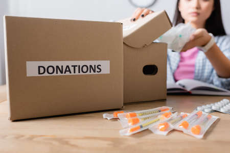 Cropped view of box with donations lettering near syringes, pills and volunteer on blurred...