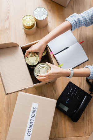 Top view of volunteer packing canned food in box near notebook and telephone on tableの写真素材