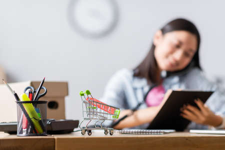 Toy shopping cart with sale lettering on price tag near stationery and volunteer talking on telephone on blurred backgroundの写真素材