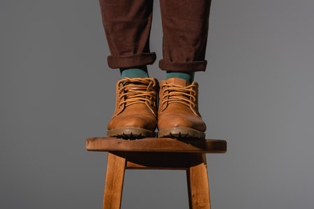 cropped view of man in boots standing on wooden chair isolated on gray