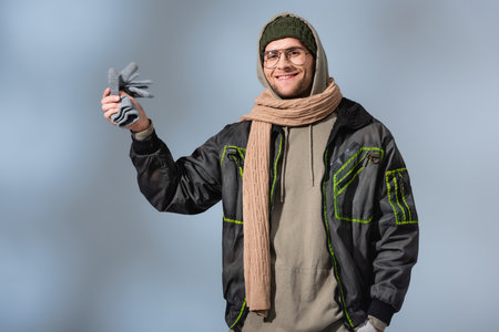 cheerful man in parka holding gloves and smiling on gray