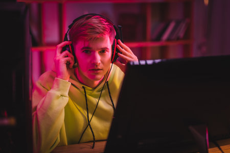 Young gamer holding headphones and looking at camera near computer monitor on blurred foreground