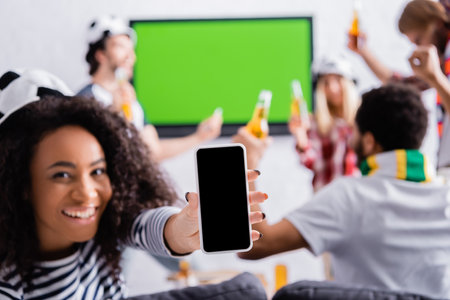 happy african american woman showing smartphone with blank screen near multicultural football fans...