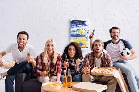 cheerful multiethnic friends watching football championship with beer, pizza and snacks at homeの写真素材