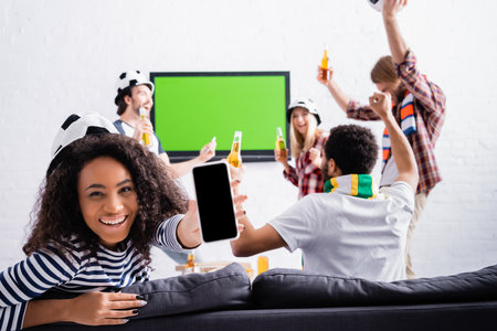 cheerful african american woman showing smartphone with blank screen near multicultural friends in football fans hats on blurred backgroundの写真素材