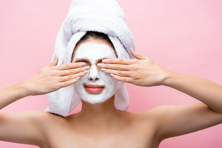 beautiful woman with towel on head and clay mask on face covering eyes isolated on pinkの写真素材