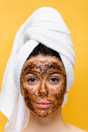 beautiful woman with towel on head and coffee mask on face isolated on yellowの写真素材