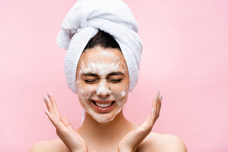 smiling beautiful woman with towel on hair and foam on face isolated on pinkの写真素材