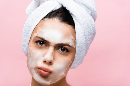 beautiful woman with towel on hair and foam on face grimacing isolated on pinkの写真素材