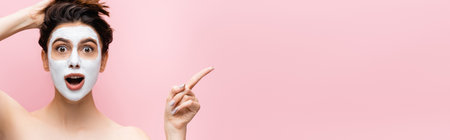 shocked beautiful woman with clay mask on face pointing aside isolated on pinkの写真素材
