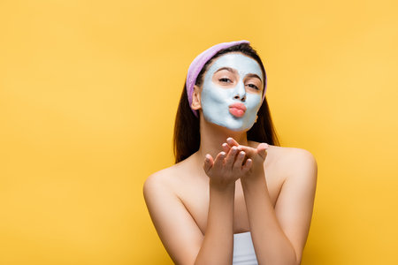 beautiful woman with clay mask on face blowing kiss isolated on yellowの写真素材