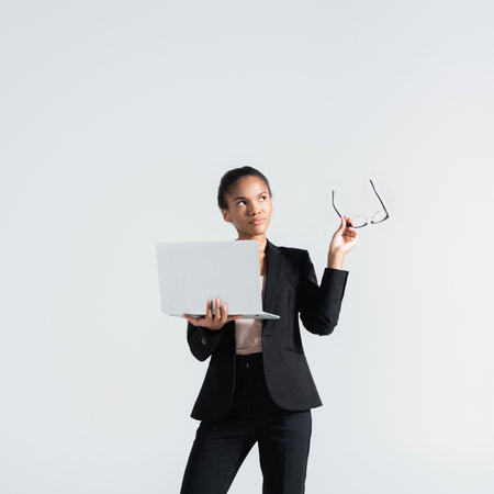 pensive african american businesswoman with glasses and laptop isolated on grayの写真素材
