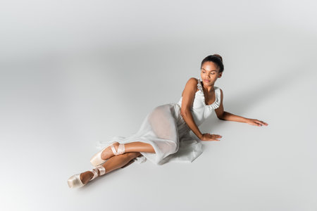 graceful african american ballerina in dress lying on floor on white backgroundの写真素材