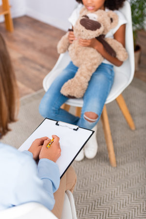 Cropped view of psychologist writing on clipboard during consultation with blurred african american girl on backgroundの写真素材