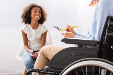 Psychologist writing on clipboard while sitting in wheelchair with blurred happy african american girl on backgroundの写真素材
