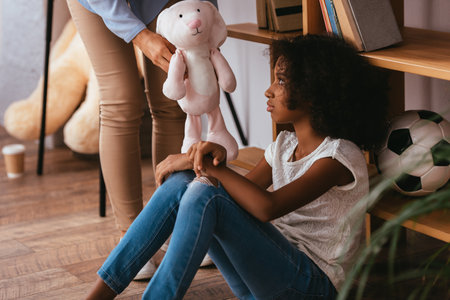 Upset african american girl with autism looking at soft toy in hands of psychologist standing near on blurred foregroundの写真素材