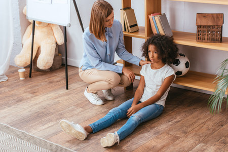 Psychologist touching shoulder of upset african american girl sitting on floor in officeの写真素材