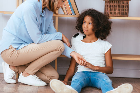 Psychologist holding hands with upset african american girl with autism sitting on floor near shelvesの写真素材