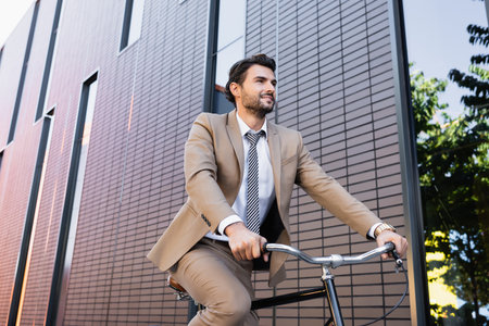 low angle view of bearded man in suit riding bicycle, smiling and looking away near buildingの写真素材
