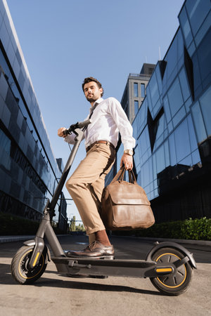 low angle view of bearded businessman in formal wear holding leather bag and standing near electric scooterの写真素材