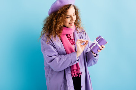 happy woman in winter coat, beret and pink knitted scarf untying ribbon on christmas present on blueの写真素材