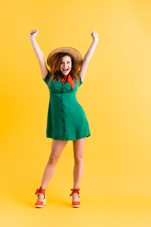 full length of amazed woman in green dress and straw hat standing with hands above head on yellowの写真素材