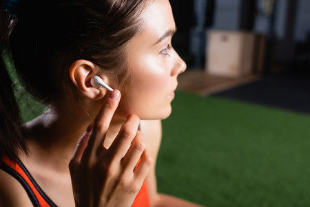 young sportswoman touching wireless earphone and looking away in sports centerの写真素材