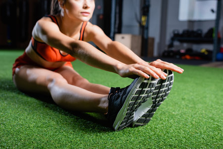 cropped view of sportswoman touching sneakers while doing seated forward bend exercise in gym,...