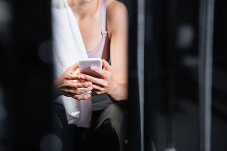 cropped view of young sportswoman messaging on smartphone while resting in gym on blurred foregroundの写真素材