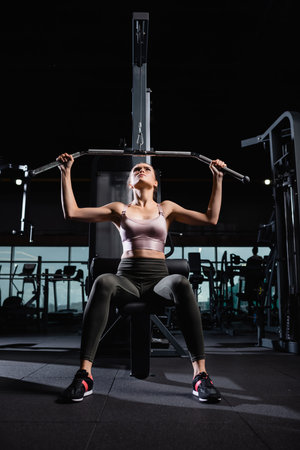sportswoman in top and leggings working out on lat machine in sports center