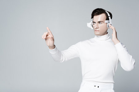 Cyborg man in headphones touching something and looking away isolated on gray