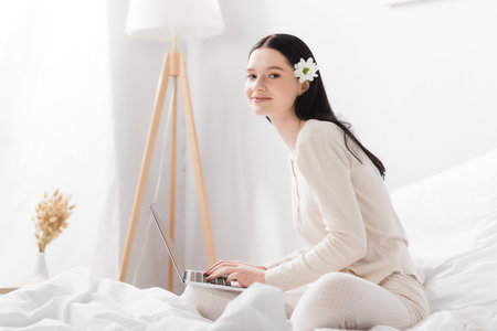 happy woman with vitiligo and flower in hair typing on laptop in bedroomの写真素材
