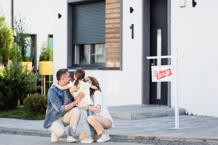 Happy parents squatting and hugging daughter near house on blurred backgroundの写真素材