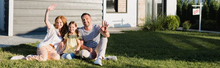 Happy parents with waving hands and daughter sitting on lawn and looking at camera near house,...
