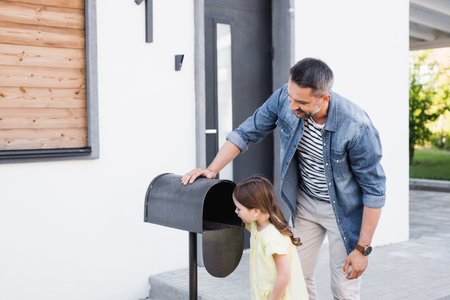 Smiling father standing near daughter looking in empty mailbox near houseの写真素材