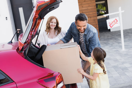 Smiling father looking at daughter while taking carton box from car trunk near wife with blurred...
