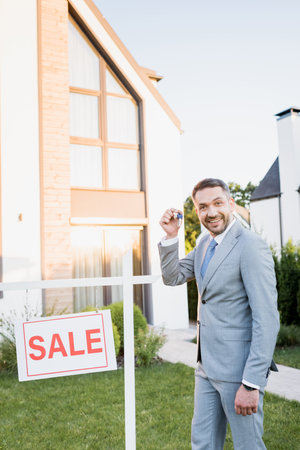 Happy broker with key looking at camera while standing near sign with sale letteringの写真素材