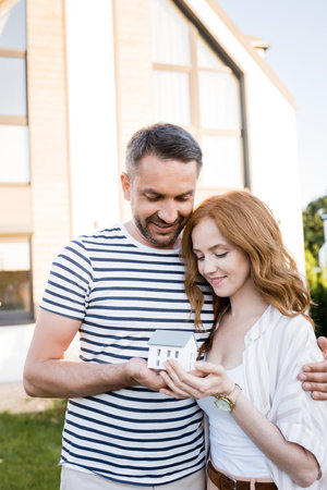 Smiling couple hugging and looking at statuette of house on blurred background