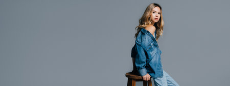 young woman in denim shirt, with naked shoulder, leaning on high chair isolated on gray, banner