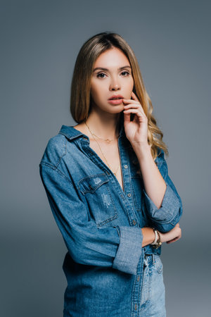 young seductive woman in denim shirt looking at camera while touching face isolated on grayの写真素材