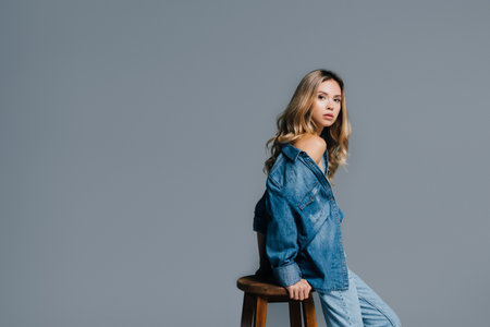 seductive woman in denim shirt, with naked shoulder, looking at camera while leaning on high stool isolated on grayの写真素材