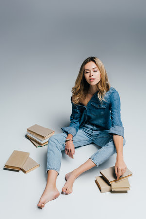 young barefoot woman in denim shirt and jeans sitting near books on grayの写真素材