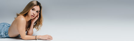 sensual topless woman in jeans touching face and looking at camera while lying on gray, bannerの写真素材
