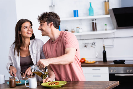 Smiling man pouring tea near girlfriend cutting waffle on kitchen tableの写真素材