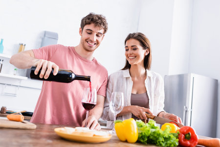 Smiling man pouring wine near girlfriend with knife and vegetables on blurred foregroundの写真素材