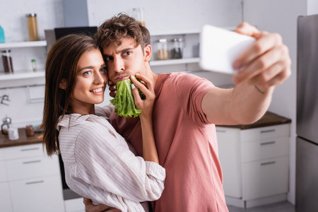 Smiling woman holding lettuce near boyfriend taking selfie on smartphone on blurred foregroundの写真素材