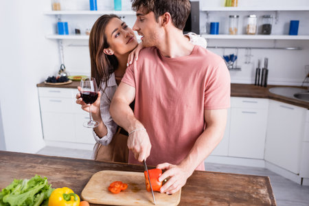Woman with glass of wine hugging boyfriend cutting paprika near vegetables on blurred foregroundの写真素材