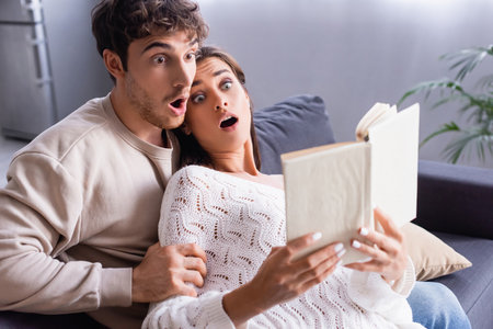 Excited couple sitting on couch while reading book on blurred foregroundの写真素材