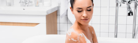 Young woman looking at wet shoulder in foam in bathtub, banner
